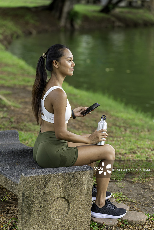 年轻亚洲女性在公园的石凳上休息，锻炼后查看手机，手里拿着一个铝制运动水瓶。图片素材