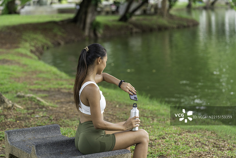 年轻亚洲女性在公园的石凳上休息，锻炼后查看智能手表，手里拿着铝制运动水瓶。图片素材