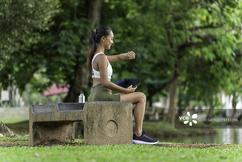 年轻亚洲女性在公园的石凳上休息，锻炼后查看她的智能手表和手机以获取重要数据。图片素材