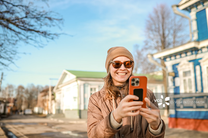 本地旅行：一位戴着帽子和太阳镜的中年女性在俄罗斯古老旅游小镇戈罗德茨的街道上使用智能手机探索。图片素材