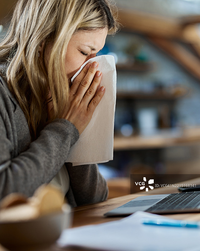 生病的女人在家用笔记本电脑工作时擤鼻子。图片素材
