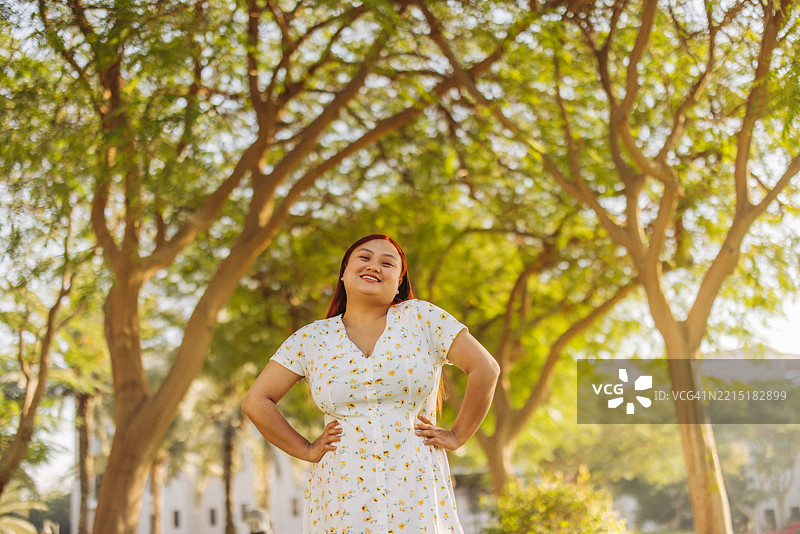 微笑的女性穿着夏季裙子，站在郁郁葱葱的树木间，沐浴在温暖的阳光下图片素材