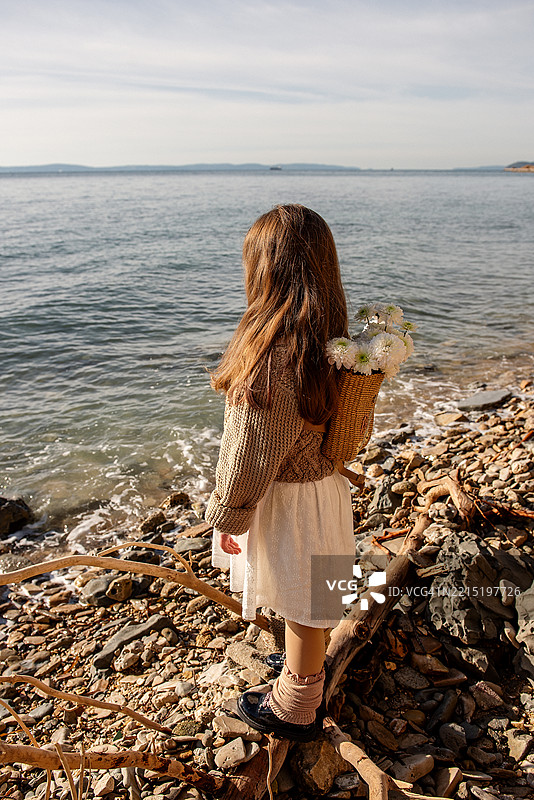 小女孩手提篮子，望着大海，篮子里装着白色花朵。图片素材
