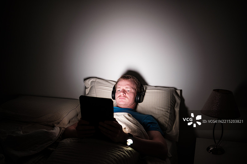 晚上躺在床上的男人使用带耳机的平板电脑图片素材