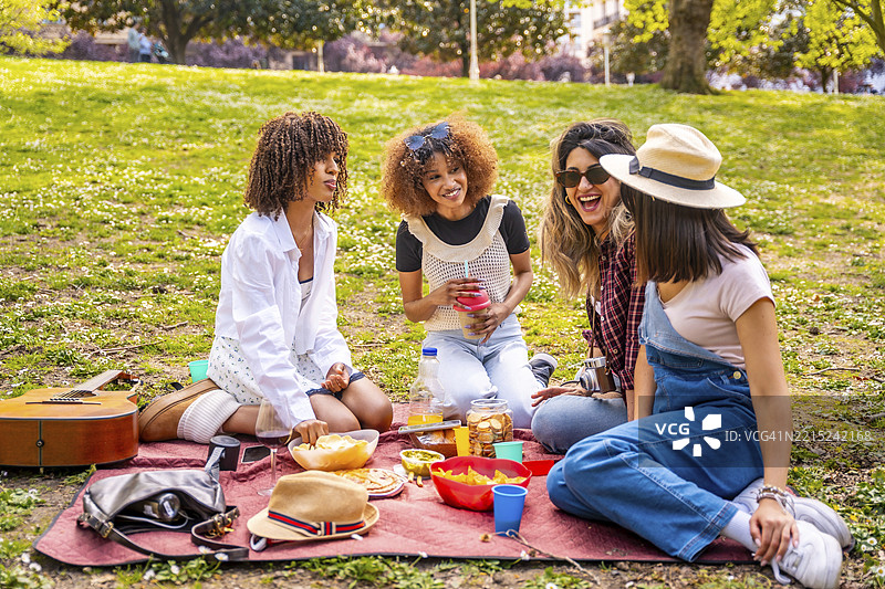 四位不同背景的女性朋友在公园享受野餐，分享食物、饮料和欢笑，阳光明媚。图片素材