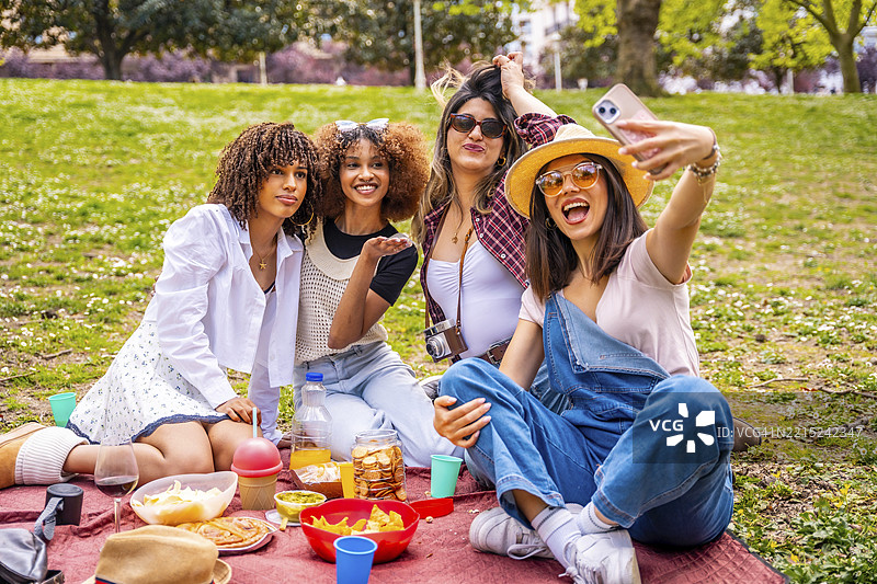 四位快乐的年轻女性在公园里享受阳光明媚的野餐，使用智能手机拍摄快乐的自拍时刻。图片素材