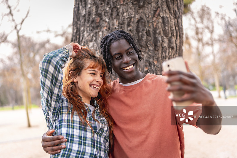 年轻的多种族情侣在公园里拥抱并用智能手机自拍，享受美好的一天。图片素材