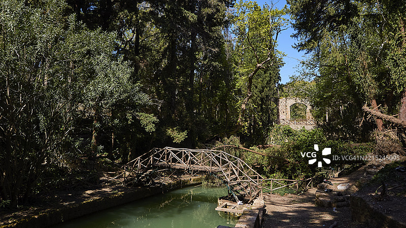 阴凉森林中的木桥横跨水面，旁边有遗迹，罗迪尼公园，城市公园，罗德镇，罗德岛，多德卡尼斯群岛，希腊，欧洲图片素材
