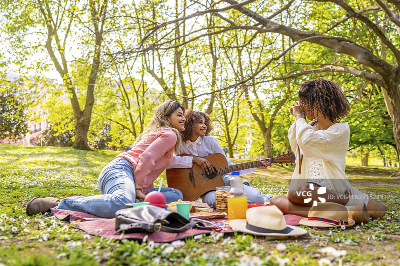 三个女性朋友在公园里享受野餐，欢笑、弹吉他、拍照，一起沐浴在阳光下。图片素材