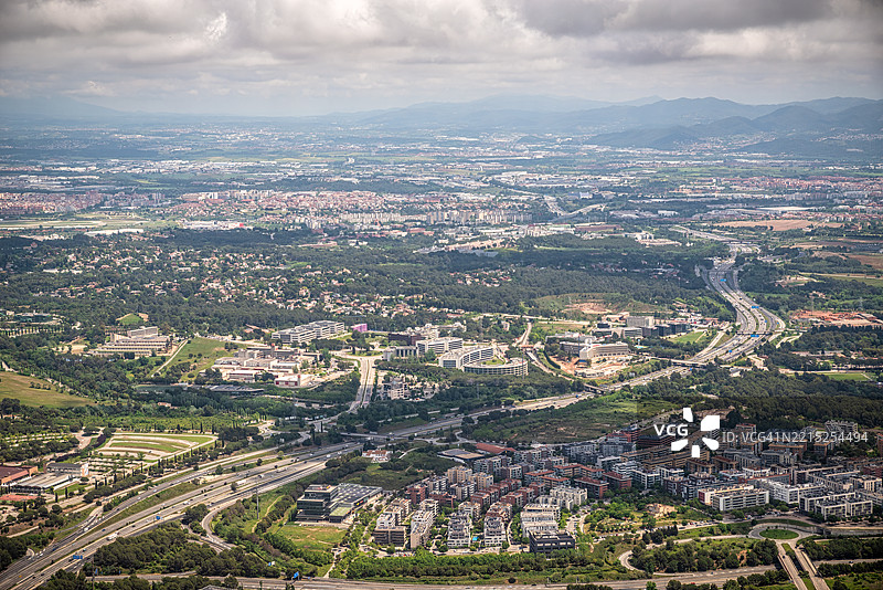 巴萨罗那省加泰罗尼亚的瓦列斯·奥西登特地区圣库加特的广阔鸟瞰图，展示了城市发展、基础设施和绿地的混合，天空多云。图片素材