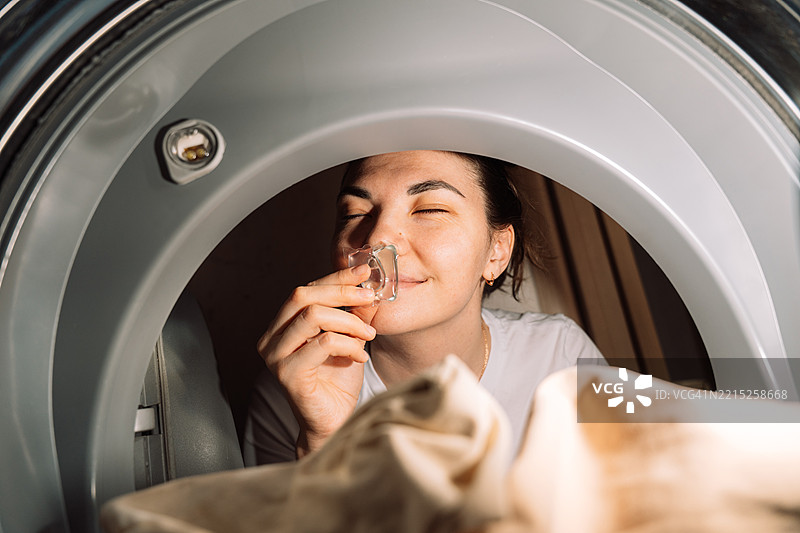 年轻女性在洗衣机里洗衣服，闻着洗衣胶囊的清香，从内部视角看，洗衣背景。图片素材