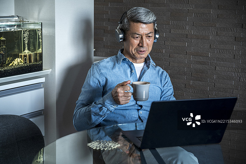 一位年长的日本男性在家中喝着咖啡，专注地看着笔记本电脑屏幕。图片素材