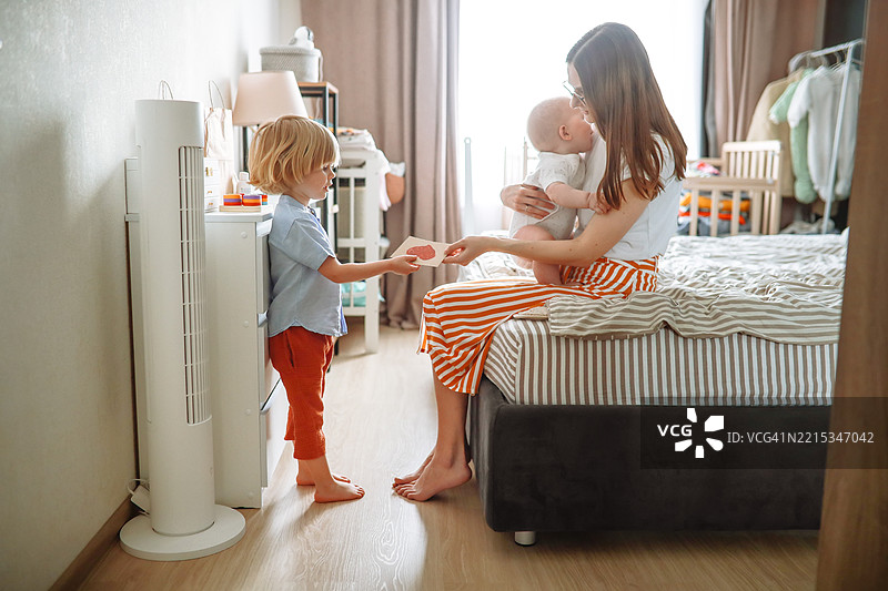 温馨的母亲节时刻：一个男孩把自己画的卡片递给抱着小妹妹的妈妈。图片素材