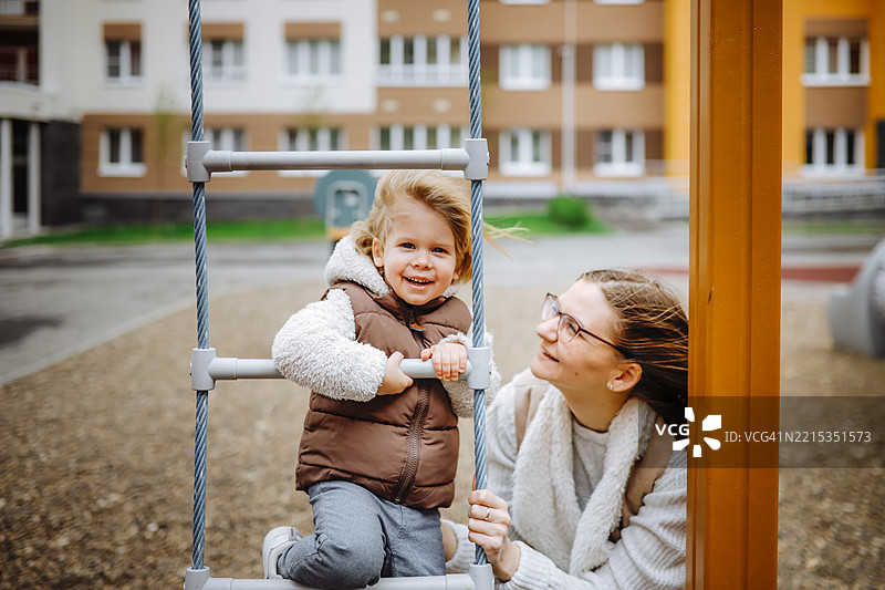 年轻母亲在高层住宅区的儿童活动区鼓励她的小孩攀爬梯子，面向镜头。图片素材