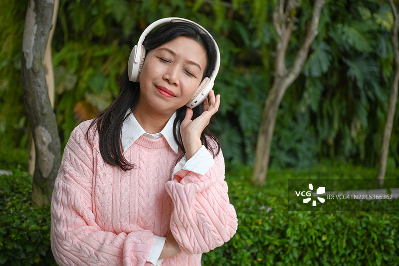 美丽的亚洲女性在公园里微笑放松，戴着蓝牙立体声耳机听音乐。图片素材