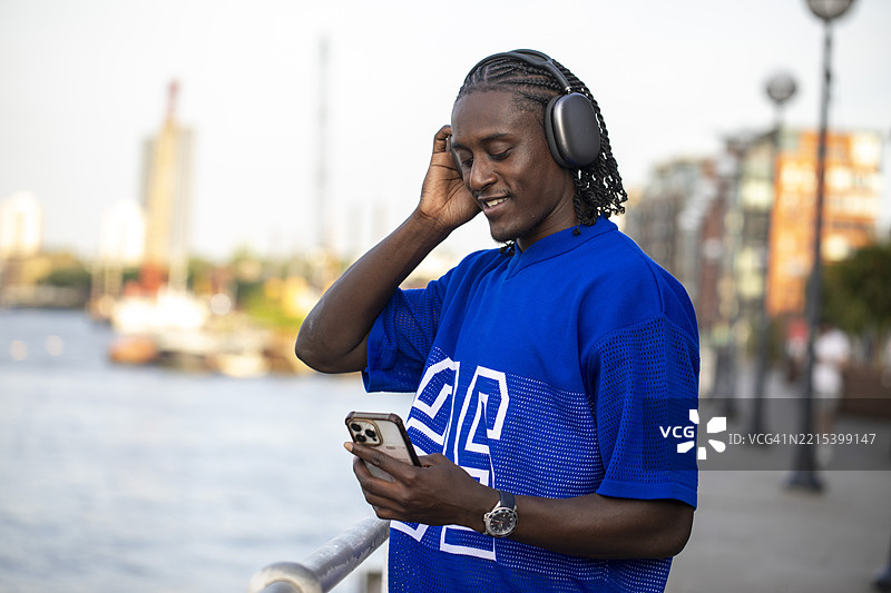 一位年轻男子在黄昏时分坐在河边，戴着耳机听音乐，手中拿着智能手机。图片素材