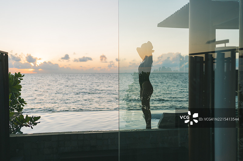 透过窗户看到年轻女性在日落时分的无边泳池边缘图片素材