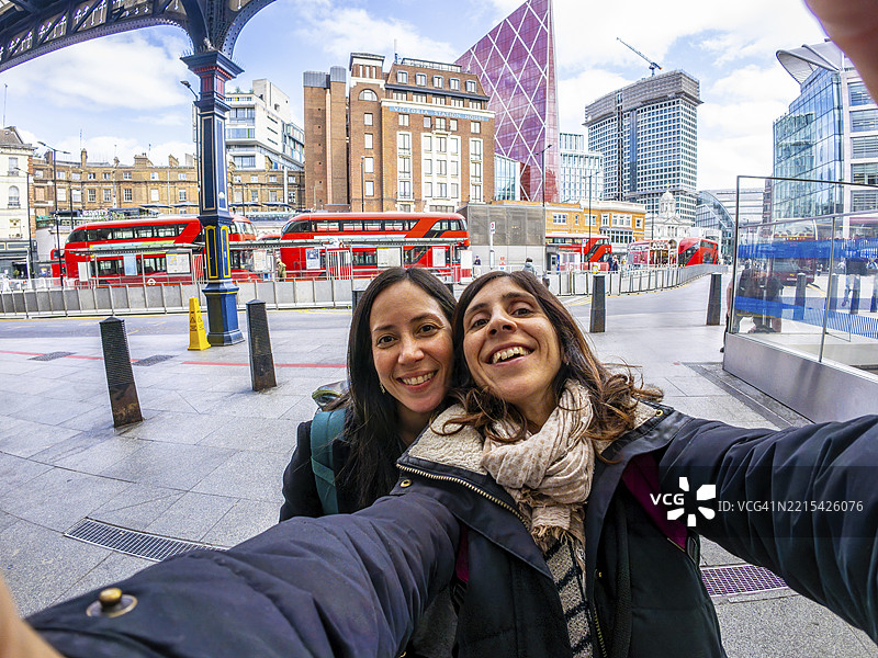 两位快乐的游客在伦敦市拍自拍，背景是红色公交车和现代建筑。图片素材