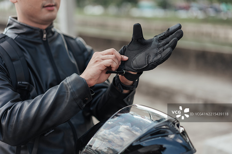 亚洲男性摩托车手在骑行前佩戴保护手套的特写。安全装备、负责任的行为、现代黑色皮夹克和头盔，背景为城市户外环境。图片素材