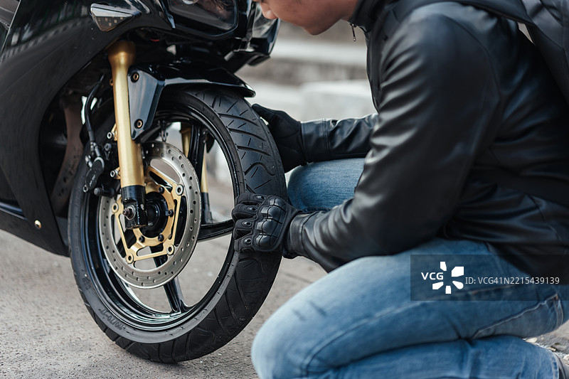 摩托车手近距离检查前轮胎状况。戴着手套的手，黑色皮夹克，运动摩托车细节。在骑行前进行安全检查，负责任的旅行准备。图片素材