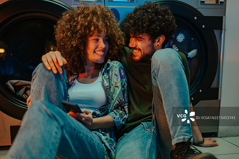 年轻情侣在自助洗衣店微笑并使用手机图片素材