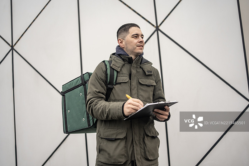 送货员手持夹板，背着绿色背包站立。图片素材