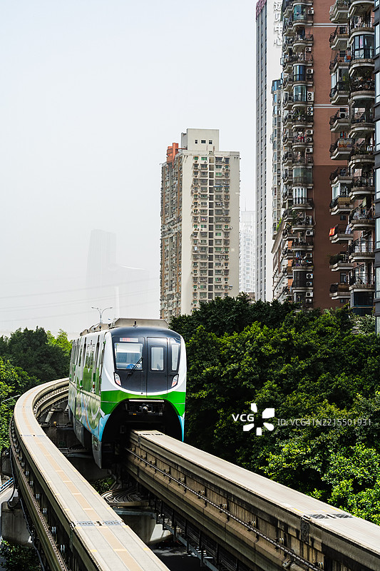 一列流线型地铁列车在繁忙的城市环境中，穿梭于高耸的住宅楼和绿树之间的高架轨道上。图片素材