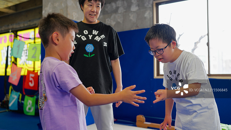 患有唐氏综合症的男孩在体育活动课堂上与朋友玩剪刀石头布，教练在一旁指导他。图片素材