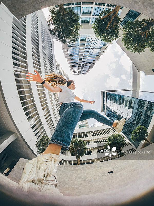 一位年轻女性从广角低视角拍摄，动态地在现代城市建筑之间跳跃。图片素材