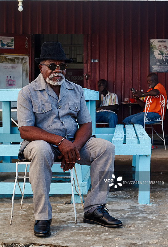 渔民村庄场景，一位穿着优雅西装和帽子的巴巴多斯男子坐在蓝色木制家具旁。六男湾，巴巴多斯，西印度群岛图片素材