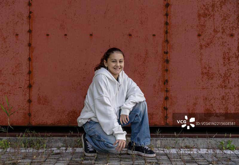 微笑的青少年女孩穿着休闲连帽衫，靠在生锈的金属墙上摆姿势图片素材