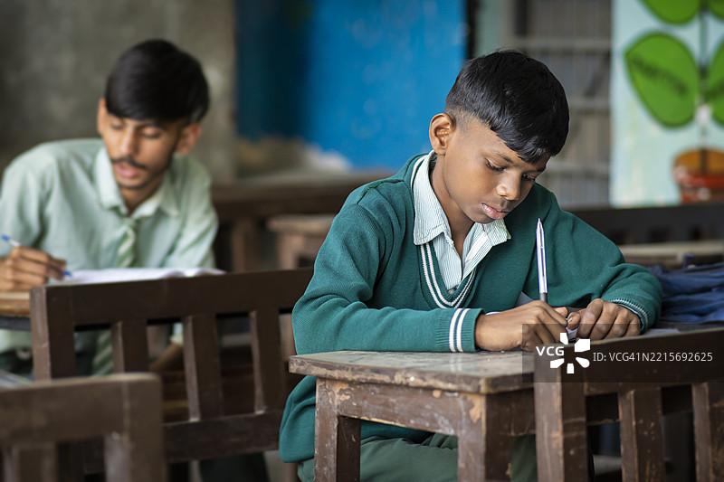 两个穿着校服的少年男生坐在教室的桌子旁，正在学习并在笔记本上写课文。图片素材