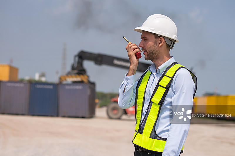 工业工程师或货运场的工人在大型集装箱和重型机械叉车旁，积极移动集装箱以管理复杂的货运操作概念。图片素材