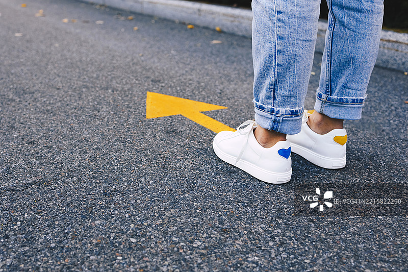 一个穿着白色运动鞋的人站在沥青路面上，面向一条大胆的黄色方向箭头。这是进步、决策和向前移动的概念。图片素材