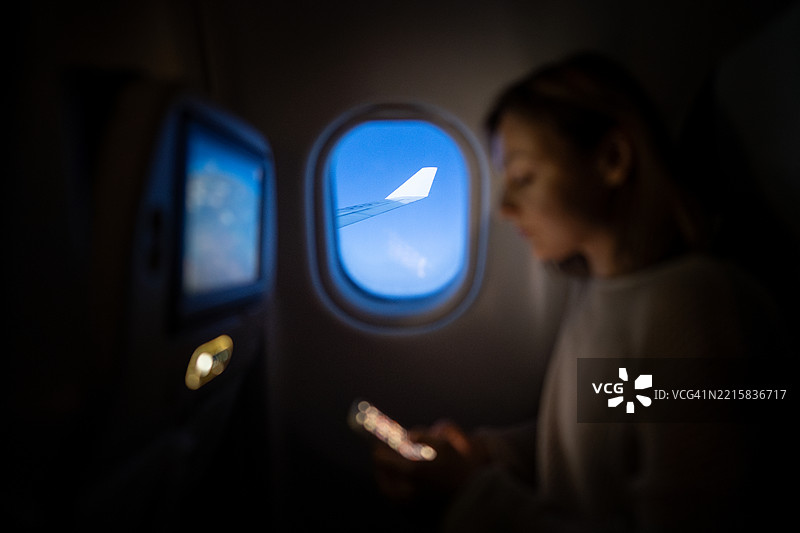 飞机窗外的机翼视角，模糊的女性正在使用智能手机。安静的经济舱旅行。图片素材