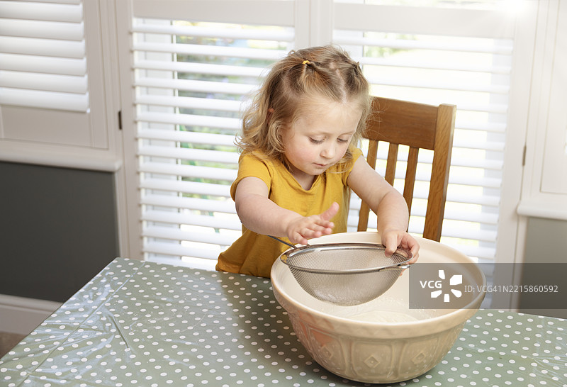 可爱的4岁女孩小心翼翼地敲打筛子，将面粉筛入厨房桌上的大搅拌碗中图片素材