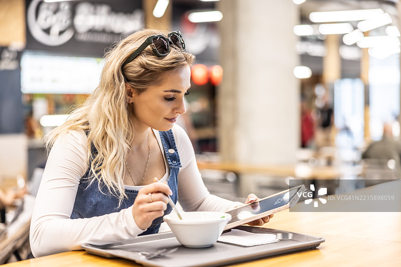 美丽的年轻女性在购物中心吃汤，面带微笑。她正在用平板电脑上网。图片素材