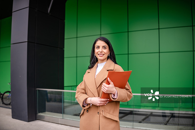 女商人在现代绿色建筑前自信地摆姿势，手中拿着一个公文包，白天的企业环境中。图片素材