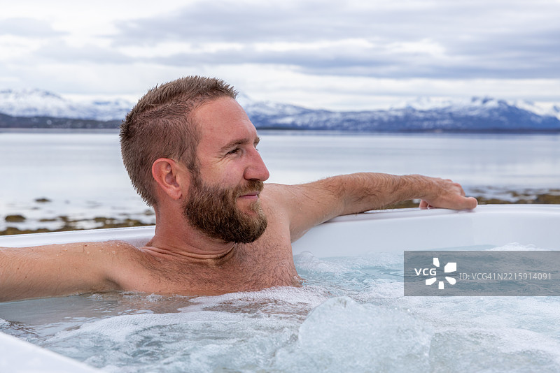 男子在户外热水浴缸中放松，俯瞰风景如画的冬季湖泊图片素材