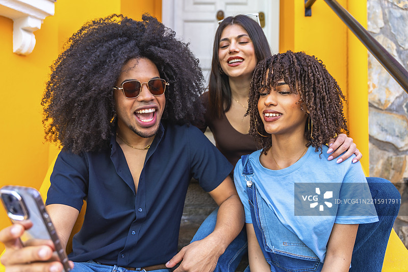 三个快乐的年轻朋友坐在色彩斑斓的建筑楼梯上，用智能手机拍自拍。图片素材