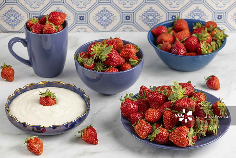 新鲜草莓摆放在蓝色陶器中，旁边是一碗酸奶，放在大理石盘子上。图片素材