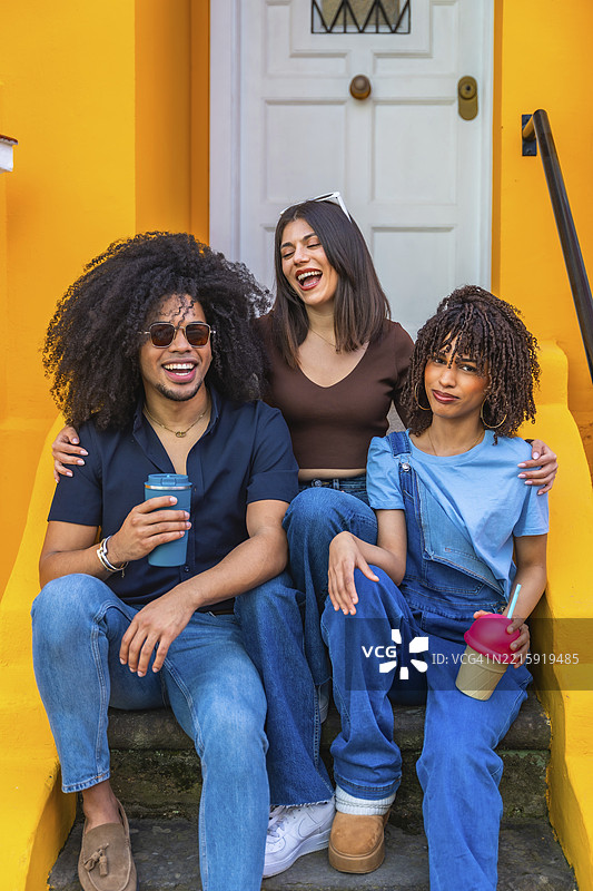 三个年轻朋友在台阶上享受轻松的时光，欢笑着，手里拿着可重复使用的杯子。图片素材