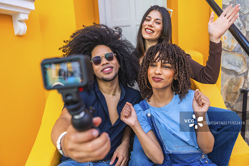 三个快乐的多民族网红坐在黄色台阶上，用运动相机拍摄视频博客图片素材