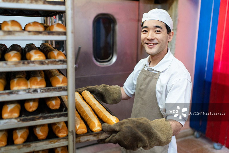 快乐的面包师傅从烘焙架上取出新鲜出炉的面包托盘图片素材