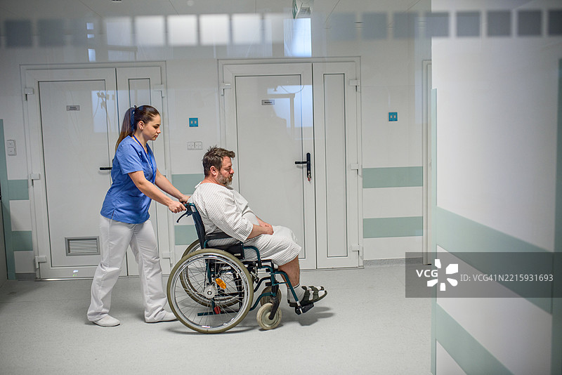 护士从侧面推着病人坐在轮椅上穿过医院走廊。图片素材
