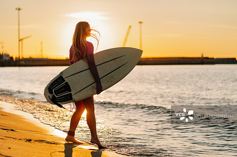 日落时分，冲浪女孩在海滩上走着，手里拿着冲浪板图片素材