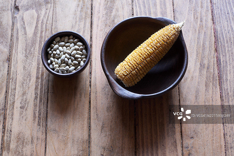 干玉米棒静静地放在装满豆子的陶瓷碗旁，摆放在风化的木质表面上，突显出乡村农业的原料。图片素材