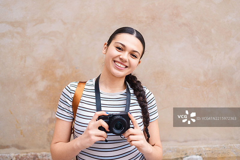 快乐的女性旅行者靠在中性色的墙上，背着背包，手持相机，表达出充满活力的旅行渴望和兴奋。图片素材