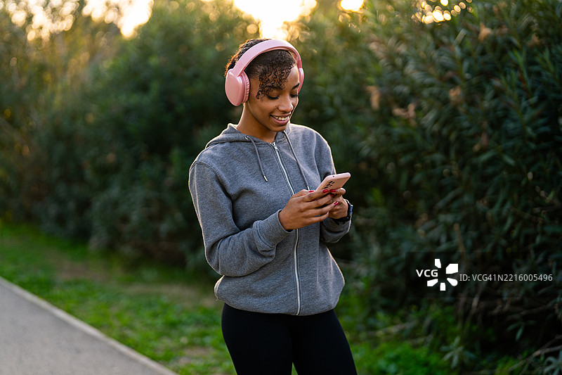 年轻女性戴着粉色耳机，一边走在公园里一边使用智能手机图片素材