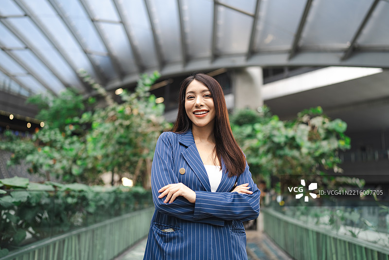 自信的年轻专业人士微笑着，双臂交叉，站在一个明亮的植物环绕的城市空间中图片素材